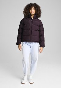 Puffersjakke i mørk lilla med høj krave og lynlås. Har en quiltet design og er parret med lyse denimjeans og hvide sneakers.