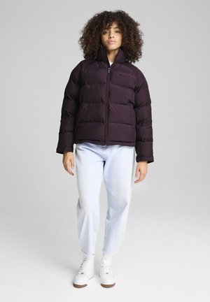 Doudoune violette foncée avec un col haut et une fermeture éclair. Présente un design matelassé et est associée à un jean en denim clair et des baskets blanches.
