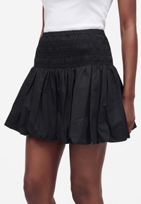 Femme portant une jupe noire courte froncée avec une taille haute froncée et un haut blanc sans manches sur un fond blanc.