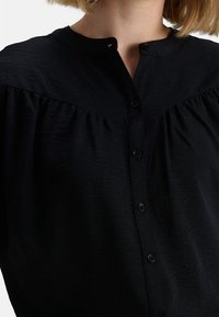 Camicia nera con colletto alla coreana, caratterizzata da un design plissettato e cinque bottoni frontali, realizzata in un morbido tessuto texturizzato.