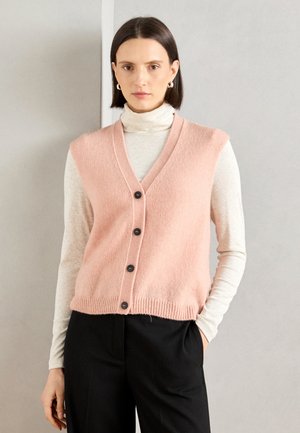 Lys rosa strikket vest med V-hals, med fem mørke knapper, ribbestrikket kant, brukt over en kremfarget turtleneck og svarte bukser.