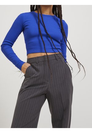 Personne avec de longues tresses portant un haut bleu court à manches longues et un pantalon gris à rayures taille haute, les mains dans les poches.