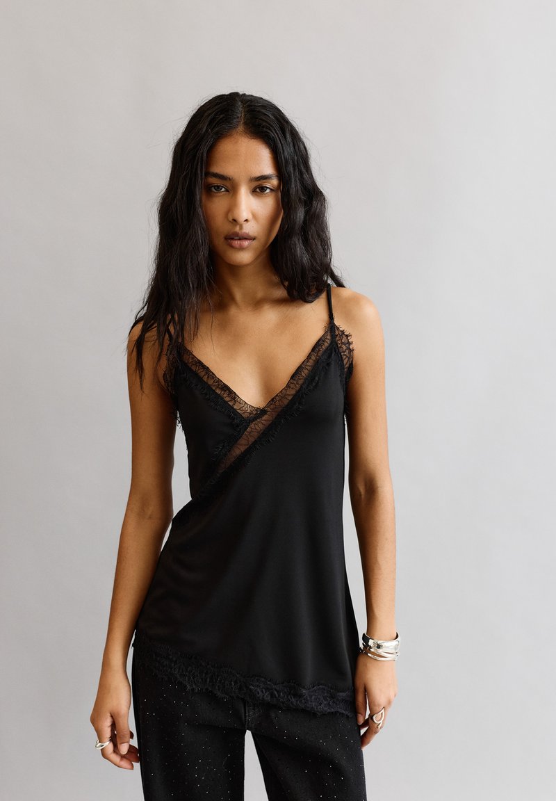 Hosszú fekete hajú nő fekete ujjatlan felsőben csipke díszítéssel, fekete nadrágban, ezüst karkötőkben és gyűrűkben, egy sima háttér előtt áll.