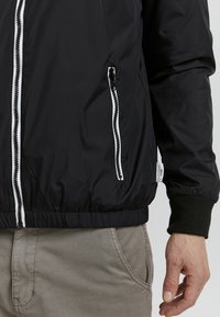 Giacca nera imbottita con dettagli della cerniera bianca, polsini elasticizzati e una tasca laterale. Texture liscia con un leggero finish lucido.