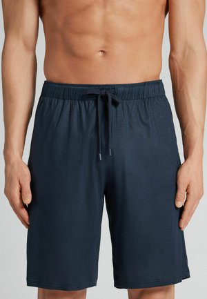 Torse d'homme portant un short bleu marine à cordon, les mains détendues le long du corps, sur un fond gris clair neutre.