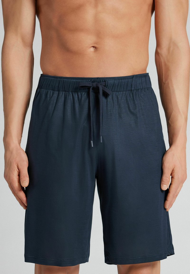 Mandetorso iført marineblå snørrebukser, hænder afslappet ned langs siderne, på en neutral lysegrå baggrund.