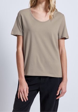 Frau trägt ein schlichtes taupefarbenes T-Shirt mit kurzen Ärmeln und Rundhalsausschnitt sowie schwarze Hose, steht mit entspannten Armen an den Seiten vor hellem Hintergrund.