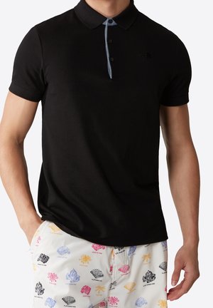 Mann trägt ein schwarzes Poloshirt und weiße Shorts mit bunten Lagerfeuersymbolen und Textmustern, steht mit einer Hand in der Tasche.