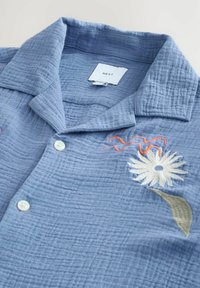 Hellblaues Hemd aus strukturiertem Stoff, mit einem Kentkragen, Knopfverschluss und floraler Stickerei in Weiß, Grün und Orange.