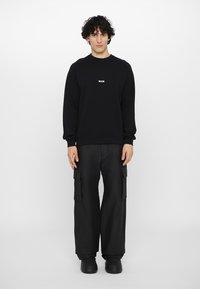 Sweatshirt noir avec un petit logo blanc sur la poitrine, associé à un pantalon cargo noir à jambes larges et des chaussures noires à enfiler.