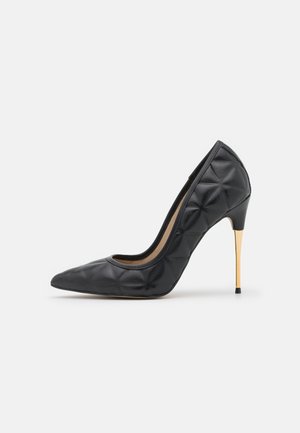 Schwarzer, gesteppter Leder-Pumps mit Absatz, spitzer Schuhspitze und schlankem, goldfarbenem, metallischem Stilettoabsatz auf weißem Hintergrund.