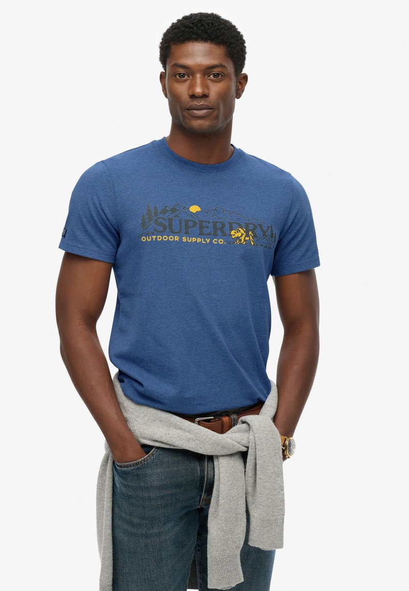Mand iført en blå Superdry Outdoor Supply Co. t-shirt, blå jeans, grå sweater bundet om livet og et guld armbåndsur, står med hænderne i lommerne.