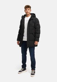Man som står klädd i en svart vadderad jacka, vit sweatshirt, mörkblå jeans och svartvita sneakers på en enfärgad ljus bakgrund.