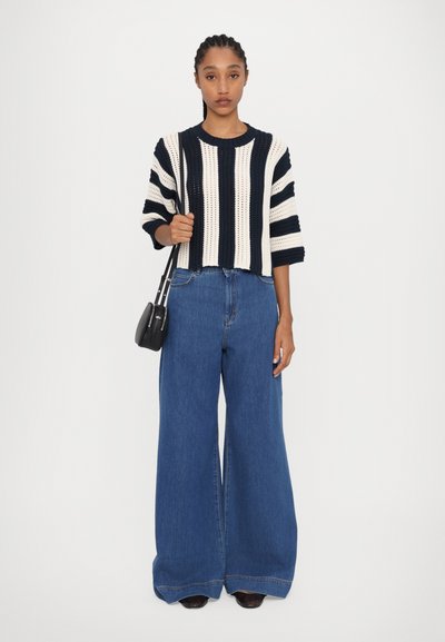 Pulover tricotat în dungi, de culoare bleumarin și crem, asociat cu blugi largi din denim albastru. Are un croi scurt și o geantă neagră mică tip crossbody.