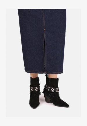 Stivaletti alla caviglia in suede nero con punta affusolata, impreziositi da una fascia geometrica in argento e frange, indossati con una gonna di jeans scura con spacco frontale.