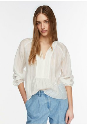 Junge Frau mit langen Haaren, die eine transparente weiße Bluse mit Puffärmeln und hellblaue hoch taillierte Jeans trägt, steht mit einer Hand in der Tasche.