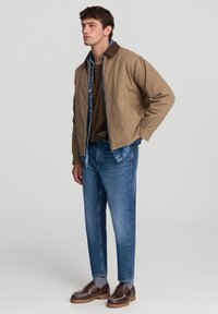 Mężczyzna w brązowej kurtce noszący niebieską koszulę dżinsową i brązowy sweter, w niebieskich dżinsach, szarych skarpetach i brązowych skórzanych butach, stojący z jedną ręką w kieszeni.