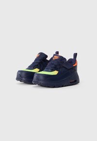 Pár modro-navy a neonovo zelených tenisk Nike s oranžovými akcentmi, šnúrkami a mäkkou podrážkou, umiestnené vedľa seba na bielom pozadí.