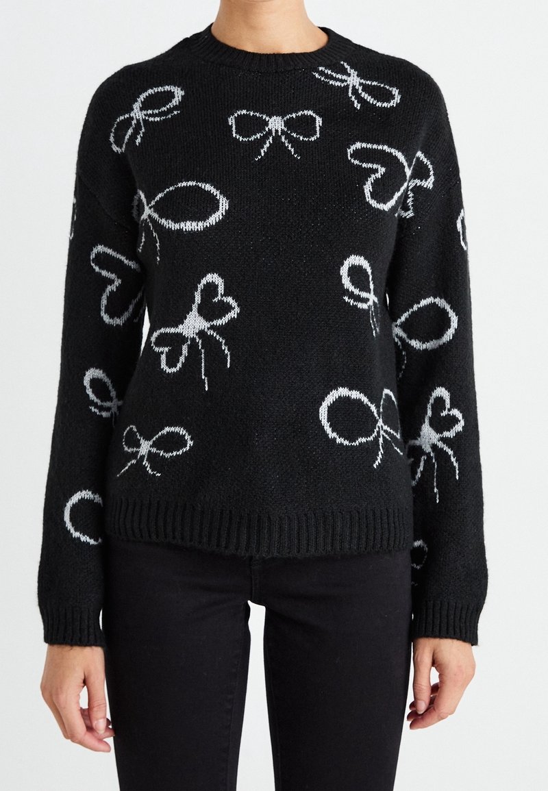 Pull en maille noire avec des motifs de nœuds blancs porté par une personne en pantalon noir sur un fond uni.