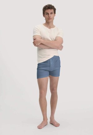 Junger Mann steht barfuß mit verschränkten Armen, trägt ein cremefarbenes Kurzarmhemd und blaue Boxershorts vor einem einfarbigen Hintergrund.