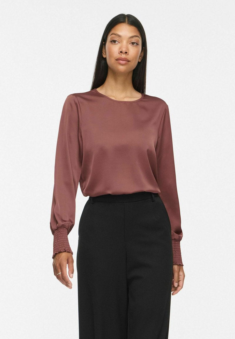 Femme portant un chemisier en satin mauve à manches longues avec des poignets smockés et un pantalon noir taille haute, debout devant un fond uni.