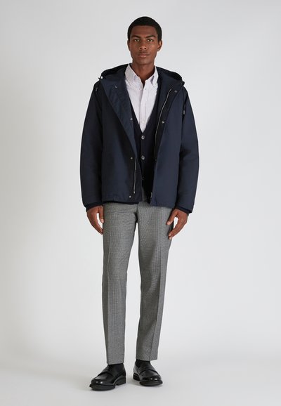 Doudoune navy avec capuche, associée à une chemise rayée blanche et bleue et un cardigan navy, un pantalon gris à motifs et des chaussures noires.