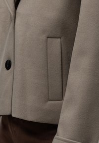 Gros plan sur le détail de la poche d’un manteau en laine beige avec un seul bouton noir et un poignet, porté sur un pantalon marron foncé.