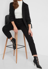 Blazer noir sur un haut blanc, associé à un pantalon noir ajusté et des bottines. Les bottines présentent un détail lacé et un bout pointu. Assis sur un tabouret.