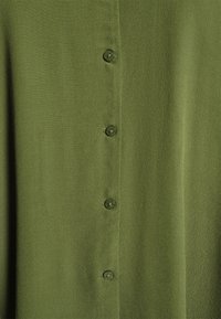 Grøn bluse med en glat tekstur, knaplukning foran med seks gennemsigtige knapper, og et enkelt, udekoreret design.