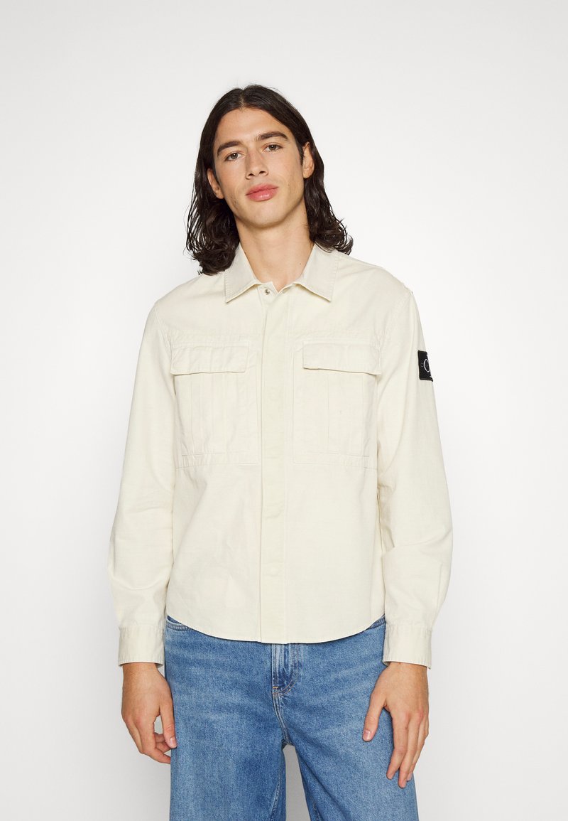 Camicia button-up beige chiaro con colletto, due tasche sul petto e una patch nera sulla manica sinistra. Indossata con jeans blu.