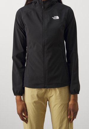 Personne portant une veste à capuche noire zippée avec le logo The North Face sur la poitrine et un pantalon beige, debout devant un fond uni.