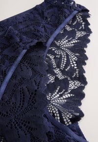 Tamsiai mėlynas nėrinių audinys su sudėtingais gėlių raštais, banguotais kraštais ir lygiu kaspino apvadu palei iškirptę ir kraštą.