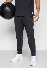 Μαύρο αθλητικό παντελόνι με στενή εφαρμογή, με πλαϊνές τσέπες και ένα μικρό λογότυπο της Nike. Συνδυασμένο με λευκά αθλητικά παπούτσια και μια μαύρη ιατρική μπάλα.