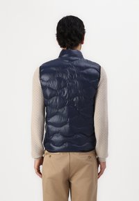 Marineblå quiltet vest med en blank finish og bølgemønster; har en ståkrage og bæres over en lys, teksturert genser.