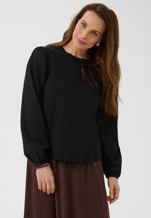 Femme aux longs cheveux bruns portant un chemisier noir à manches bouffantes ajourées et une jupe marron, debout devant un fond uni.