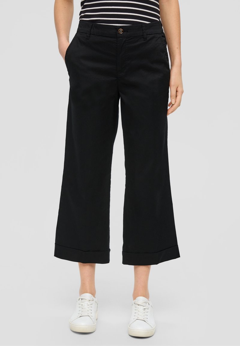 Personne portant un pantalon noir ample et court, un haut rayé et des baskets blanches, debout les mains dans les poches sur un fond uni.