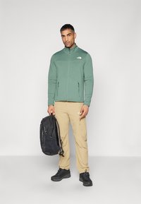 Giacca in pile verde con zip e colletto alto, pantaloni beige, zaino nero e scarpe nere. Presenta il logo sulla giacca e tasche con zip.