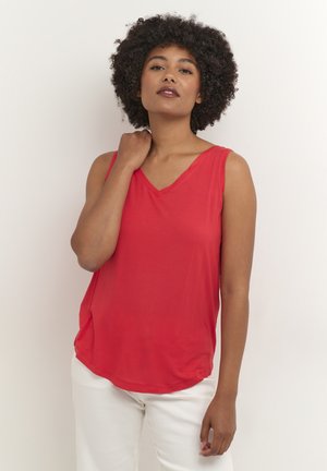 Haut sans manches rouge en tissu léger avec un décolleté en V. Ourlet arrondi avec fentes sur les côtés. Le modèle est associé à un pantalon blanc, se tenant contre un fond uni.