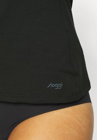 Black Sloggi top in soft fabric, featuring a rounded hem and light blue logo at the bottom left, paired with black underwear.