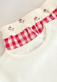 Fehér bordázott anyagú felső, smockolt piros és fehér kockás gallérral, apró hímezett virágokkal a krémszínű háttéren.