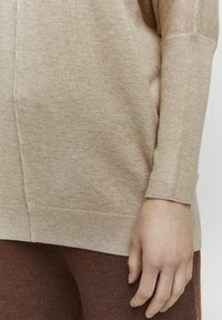 Hellbeige gestrickter Pullover mit geripptem Ausschnitt und Bündchen, in lockerer Passform. Die Textur wirkt weich und leicht meliert.