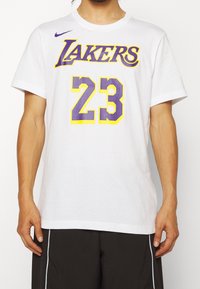Camiseta blanca de algodón con la palabra "LAKERS" en texto morado y amarillo, número "23" en morado con una textura elevada, cuello redondo y mangas cortas.