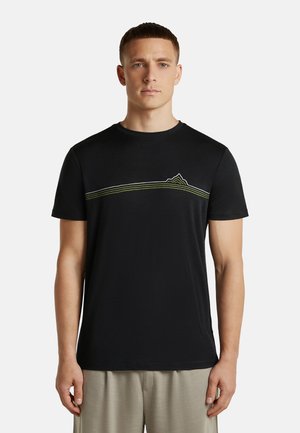 Mann trägt ein schwarzes kurzärmeliges T-Shirt mit einem dünnen horizontalen grünen Berglinien-Design über der Brust, kombiniert mit beigen Hosen.