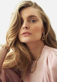 Guldig länkarmband, guldörhängen i ringform, mjuk rosa blus med puffärmar, långt vågigt hår, naturlig makeup, neutral bakgrund.