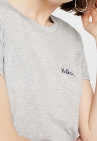 Šedé bavlněné tričko s kulatým výstřihem, krátkými rukávy a kruhovým logem v námořnické modři s nápisem "Hollister Southern California" na hrudi.