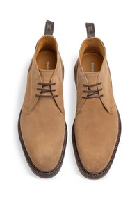 Botines de ante color tostado con un acabado suave, punta redondeada y cordones marrones en contraste. Presenta una suela cosida y tiradores para facilitar el calzado.