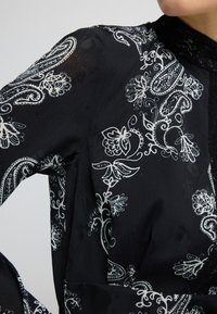 Schwarze Bluse aus Stoff mit weißen Blumen- und Paisley-Mustern, einem strukturierten Kragen und filigranen Spitzenakzenten am Ausschnitt.