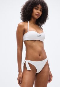 Σετ λευκού μπικίνι με τοπ μπαντό με κεντρικό λογότυπο και σλιπ με κορδόνια στο πλάι. Λείες υφές, μινιμαλιστικός σχεδιασμός και χωρίς σχέδια.