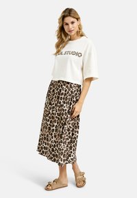 Weißes, kurzes T-Shirt mit "SUL STUDIO" im Leopardenmuster. Fließender, beige Midi-Rock mit braunen Leopardenflecken. Braune Sandalen mit Schnallen.