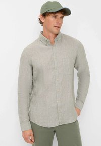 Light gray long-sleeve shirt with a button-down collar, single chest pocket, and textured fabric. Paired with a green cap.